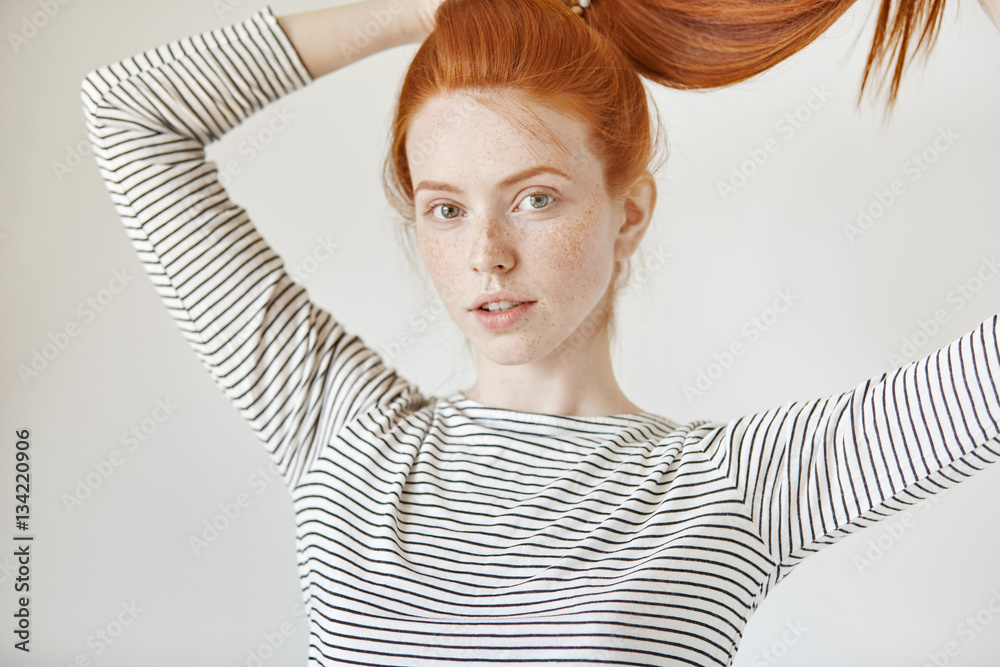 Pretty girl with green eyes and freckles smiling joyfully tying her long red  hair in ponytail,