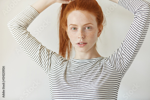 Indoor shot of beautiful freckled teenage girl with long ginger hair gathered in ponytail dressed casually posing indoors, adjusting her hairstyle before going to college. Youth and tenderness concept photo