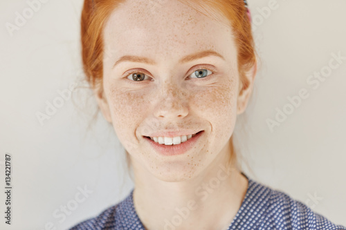 Close up portrait of beautiful young redhead model with different colored eyes and healthy clean skin with freckles smiling joyfully, showing her white teeth, posing indoors. Heterochromia in human photo