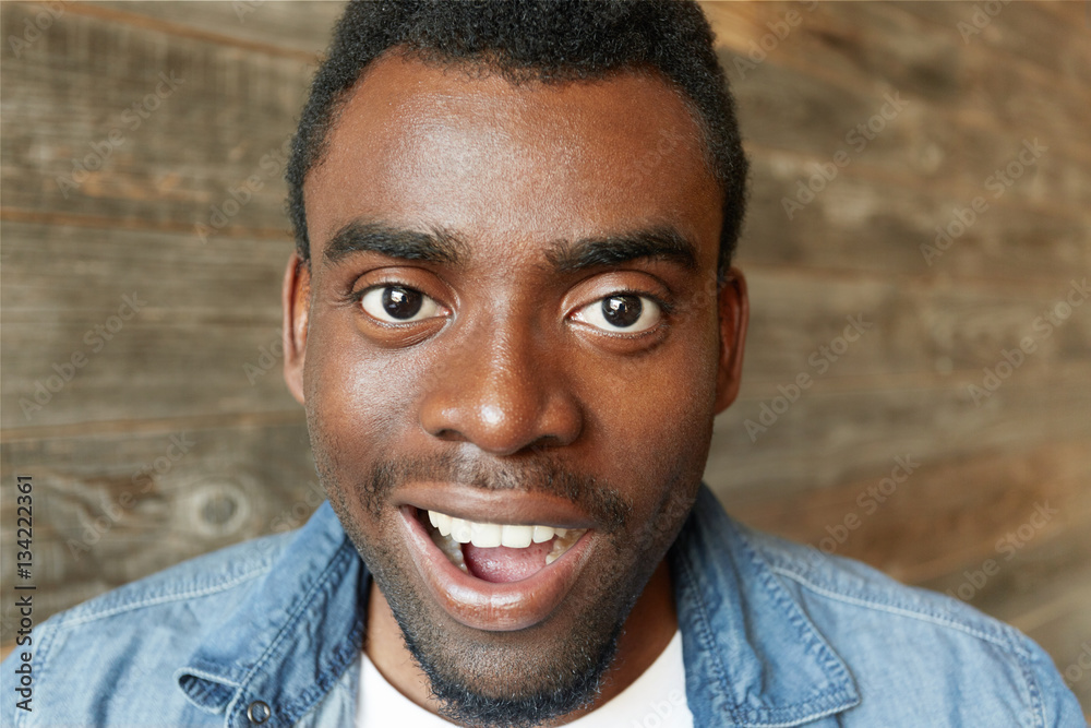 Close up portrait of happy young African male wearing white t-shirt and denim jacket having amazed look, posing isolated against wooden wall background. Human face expressions, emotions and feelings Close up portrait of happy young African male wearing white t-shirt and denim jacket having amazed look, posing isolated against wooden wall background. Human face expressions, emotions and feelings