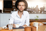 Attractive fashionable young entrepreneur with beard having coffee at modern cafe, looking serious while waiting for his partner to join him for lunch. Caucasian man in headwear relaxing indoors alone