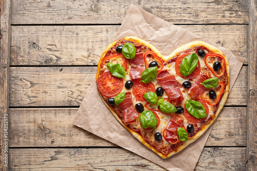 Pizza heart shaped love concept Valentine's Day symbol restaurant romantic dinner food. Prosciutto, olives, tomatoes, basil and mozzarella cheese baked meal on vintage wooden table background.