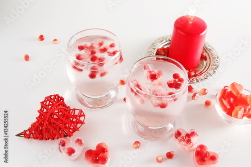 Romantic still life. Two glasses with alcoholic cocktail and ice, decorated with pomegranate, red candle, handmade heart, jelly candies, confetti. Saint valentine's day celebration.