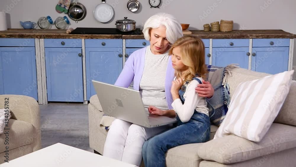 Mature grandmother and her little granddaughter sitting on sofa and looking on screen of laptop computer. Happy girl showing her granny some pictures or photos. Mature grandmother and her little granddaughter sitting on sofa and looking on screen of laptop computer. Happy girl showing her granny some pictures or photos.