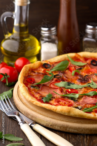 Mediterranean traditional pizza with ham, tomatoes, olives and herbs on wooden table.