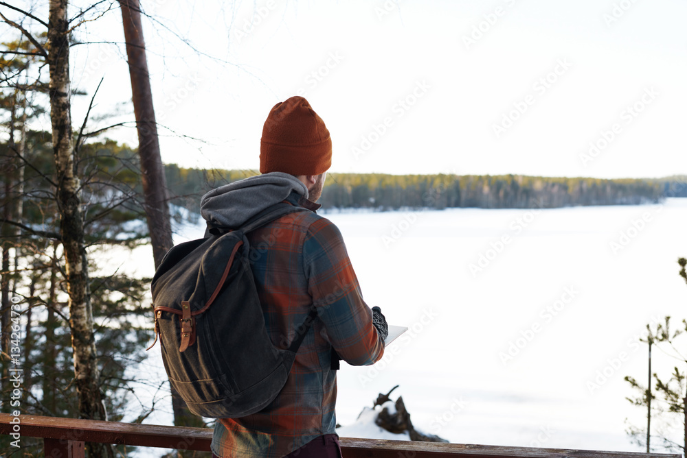 Backpacker near forest lake in winter period. Hipster dressed man write notes in travel book. Copy space for advertising text or tourist goods. Adventure concept.