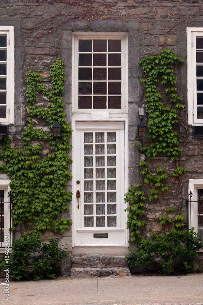 White front door and sash windows.  Green vines climbing up stone wall.  Stone front steps and sidewalk in front of quaint town house in Old Montreal, Montreal, Canada.