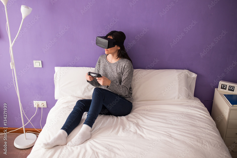 Fototapeta premium Young woman sitting on the bed at home having fun playing a virt