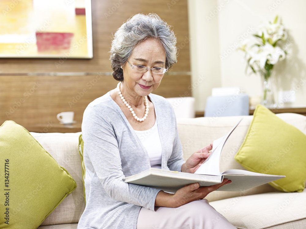 senior asian woman sitting on couch reading a book at home senior asian woman sitting on couch reading a book at home