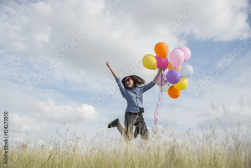 Beautiful woman wearing blue shirt,hat and glass holding colorful balloons in the meadow against blue sky and clouds,spreading hands,jump off the ground.
