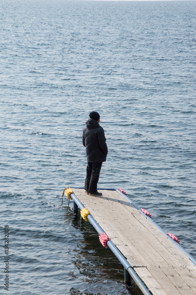 Man dressed in black with back to camera standing on a wooden dock jutting into Lake Baikal, Siberia. The water is a dark blue gray. 