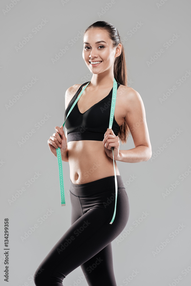 Smiling charming young sportswoman standing and holding measuring tape Smiling charming young sportswoman standing and holding measuring tape