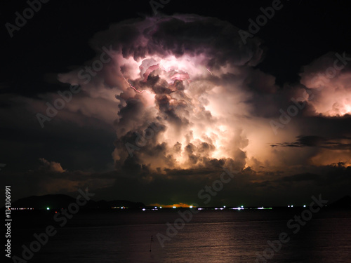 A lot of clouds in the sky with storms and lightning inside With a light from a fishing boat that is far away, Above the calm sea during the rainy season at night In the eastern part of Thailand