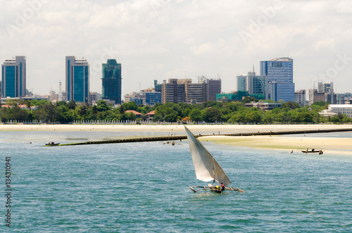 Coastline of Dar Es Salaam. A fishing boat is sailing along the beach towards the huge drainage pipe extending in to the ocean. The skyline with skyscrapers is in the background