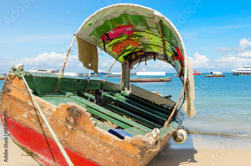Wooden old boat sitting on the beach in Zanzibar on a perfect summer day under blue sky with calm sea. Many vessels are seen in the background extending to the horizon