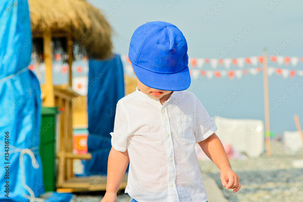 Baby boy in blue cap with sad expression. Frown baby boy on the stone beach in summer vacation