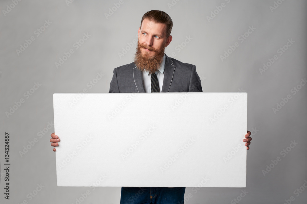 Hipster business man with beard and mustashes in suit standing holding white banner, looking away over grey background Hipster business man with beard and mustashes in suit standing holding white banner, looking away over grey background