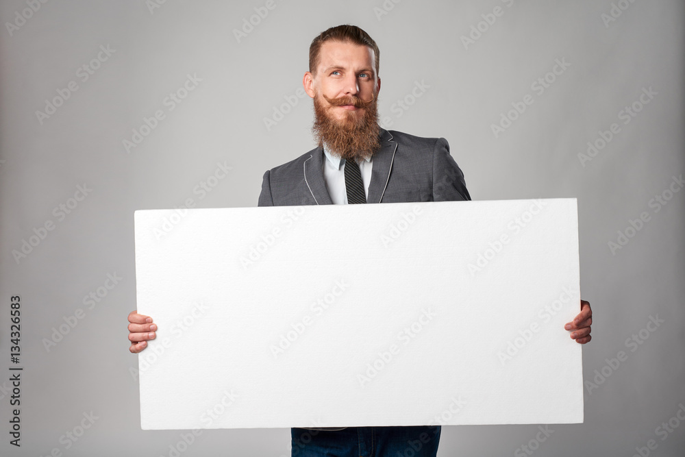 Hipster business man with beard and mustashes in suit standing holding white banner, looking up in thoughts daydreaming, over grey background Hipster business man with beard and mustashes in suit standing holding white banner, looking up in thoughts daydreaming, over grey background