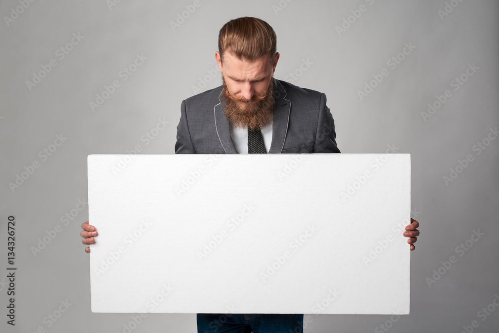 Hipster business man with beard and mustashes in suit standing holding white banner, looking down at it over grey background Hipster business man with beard and mustashes in suit standing holding white banner, looking down at it over grey background