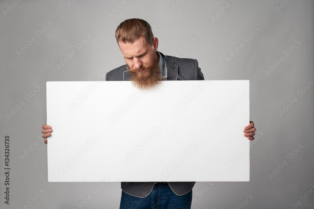 Hipster business man with beard and mustashes in suit standing holding white banner, looking down at it over grey background Hipster business man with beard and mustashes in suit standing holding white banner, looking down at it over grey background