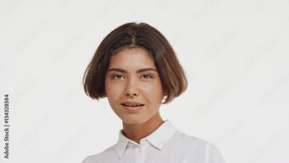 Young beautiful brunette east asian female in shirt flirting at camera smiling showing tongue waving hand on white background in slowmotion Young beautiful brunette east asian female in shirt flirting at camera smiling showing tongue waving hand on white background in slowmotion