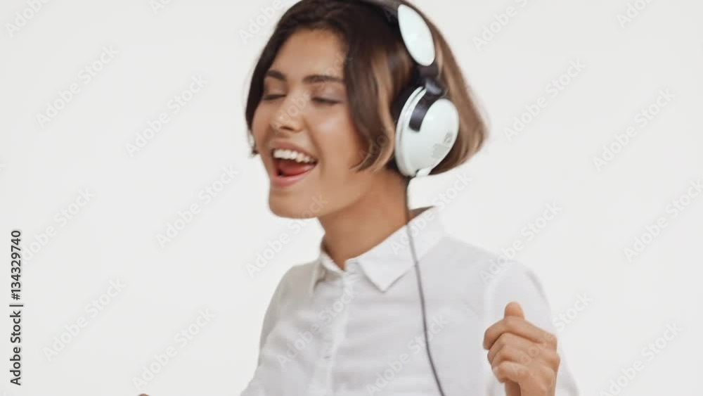 Young beautiful brunette east asian female in shirt listening music through headphones dancing smiling singing on white background in slowmotion Young beautiful brunette east asian female in shirt listening music through headphones dancing smiling singing on white background in slowmotion