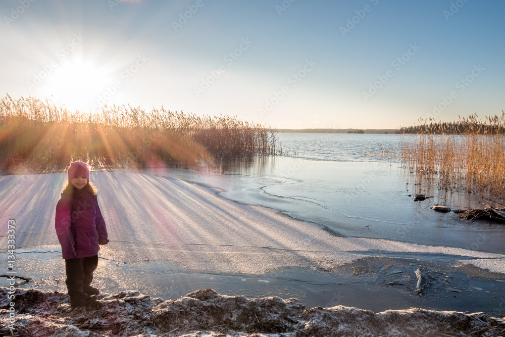 Φωτογραφία Young girl standing at a shore looking at winter sunset with sun flares on the snow and ice