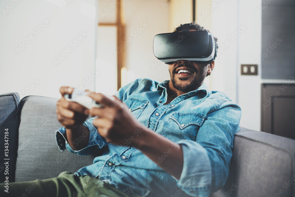 Bearded african man enjoying virtual reality glasses while sitting on  sofa.Happy young guy with vr headset or 3d spectacles and controller  gamepad playing video game at home.Blurred. Stock Photo | Adobe Stock