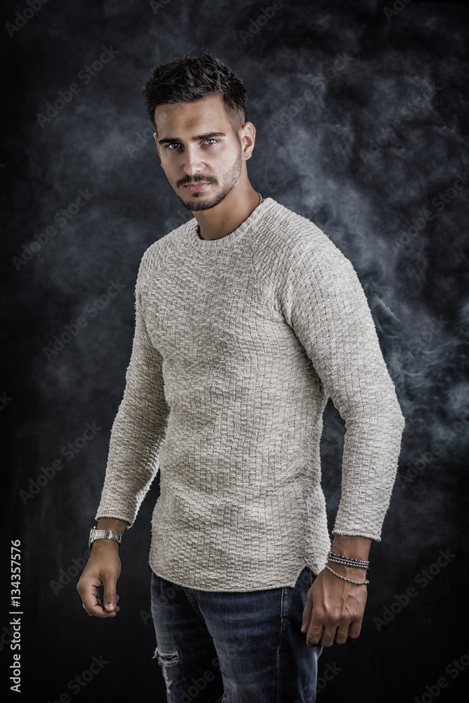 Handsome young man wearing white sweater and ripped jeans, on dark background in studio shot