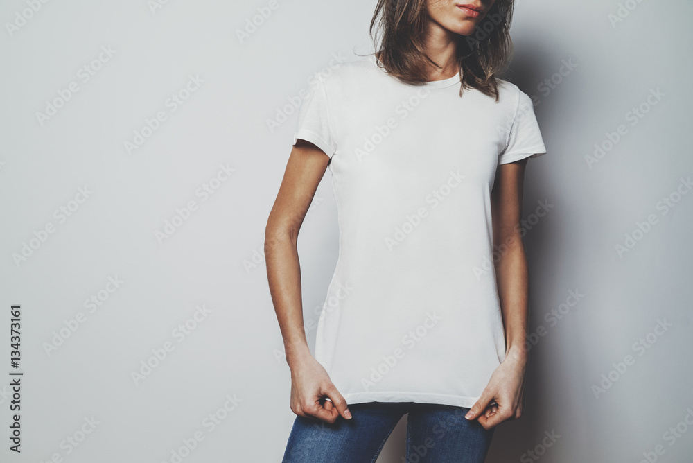 Fototapeta premium Young hipster girl wearing blue jeans and blank white t-shirt with area for your logo or design, mock-up of white t-shirt, white wall in the background