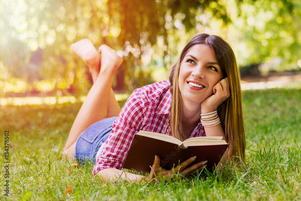 Young nice attentive woman lies on green grass and reads book against city park. Young nice attentive woman lies on green grass and reads book against city park.