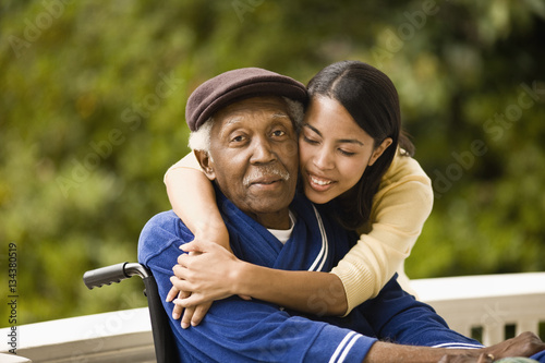 Young woman leans over and embraces her grandfather sitting in a wheelchair as they pose for a portrait on a porch.