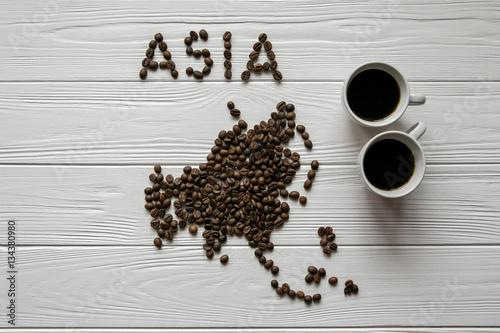 Map of the Asia made of roasted coffee beans laying on white wooden textured background with two cups of coffee