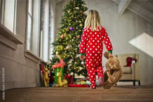 Young girl walking towards the Christmas tree with her teddy bear. photo