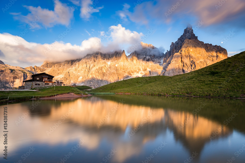 The Pale di San Martino peaks (Italian Dolomites) reflected in the water at sunset, with an alpine chalet on background.