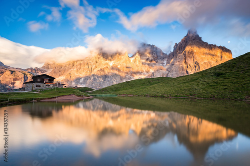Fototapeta Naklejka Na Ścianę i Meble -  The Pale di San Martino peaks (Italian Dolomites) reflected in the water at sunset, with an alpine chalet on background.