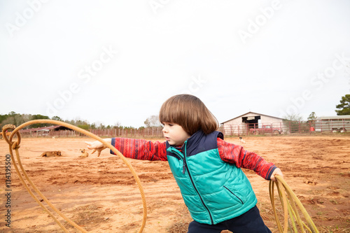 Handsome small toddler boy training making loope and throwing lasso in ranch. Red clay background, rural. Country lifestyle