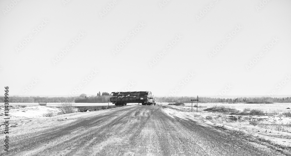 Obraz premium A semi truck and trailer loaded with round straw bales turning down a rural countryside gravel road in a black and white agricultural landscape