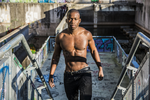 Portrait of a hot black man shirtless in urban environment, walking on metal stairs, looking at camera