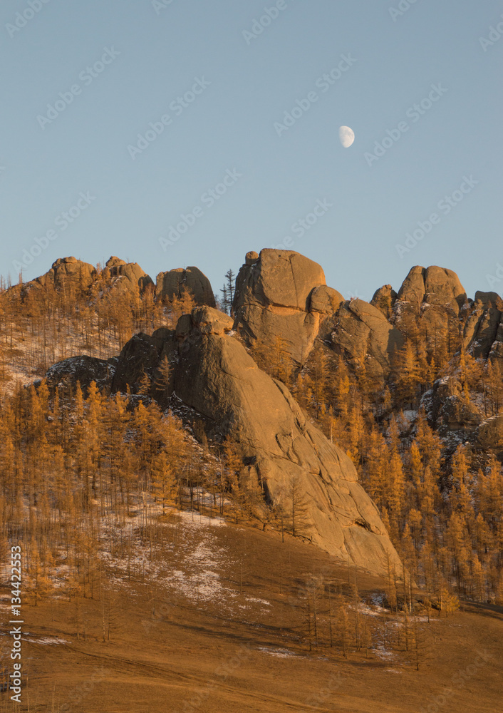 Rugged mountains with large rust colored stones with sparse vegetation. The rising moon is seen above the mountains. Photographed in Gorkhi-Terelj National Park, Mongolia.