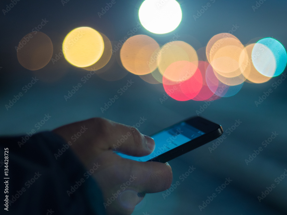 Man using smartphone at night, bokeh light in blurred background.