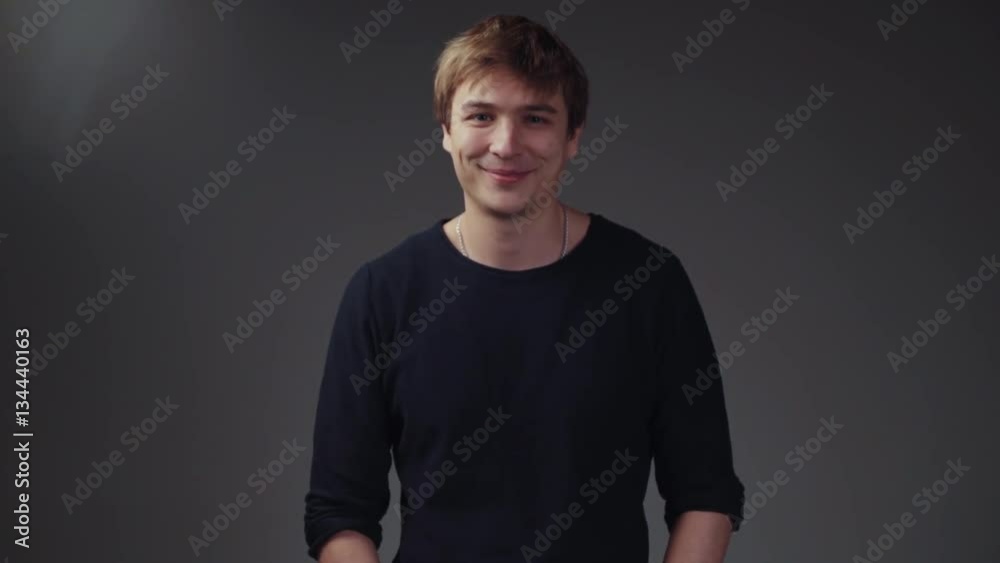 Handsome young European man in a stylish black sweater on the grey screen. Funny boy looking down, lifts his head and looks towards the camera, gives a cute playful smile.