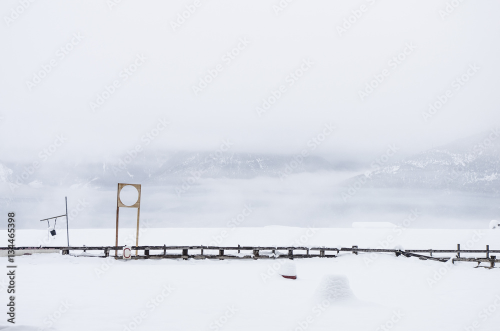 A wooden fence almost entirely covered by snow. Beyond the fence there is a valley and mountains hidden by the fog