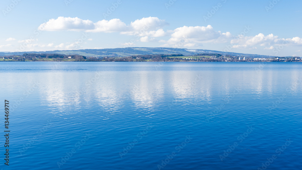 Clear day.  Lake Zug in central Switzerland. Clouds are reflected in water lake. At the edge water is clear and see the bottom of the lake.