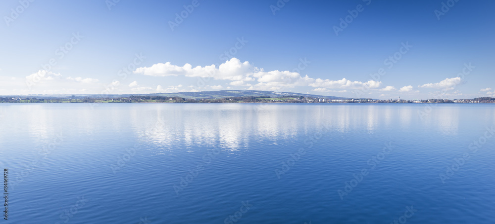 Clear day. Lake Zug in central Switzerland. Clouds are reflected in water lake. At the edge water is clear and see the bottom of the lake. Clear day. Lake Zug in central Switzerland. Clouds are reflected in water lake. At the edge water is clear and see the bottom of the lake.