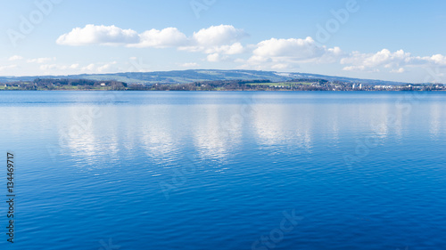 Fototapeta Naklejka Na Ścianę i Meble -  Clear day.  Lake Zug in central Switzerland. Clouds are reflected in water lake. At the edge water is clear and see the bottom of the lake.