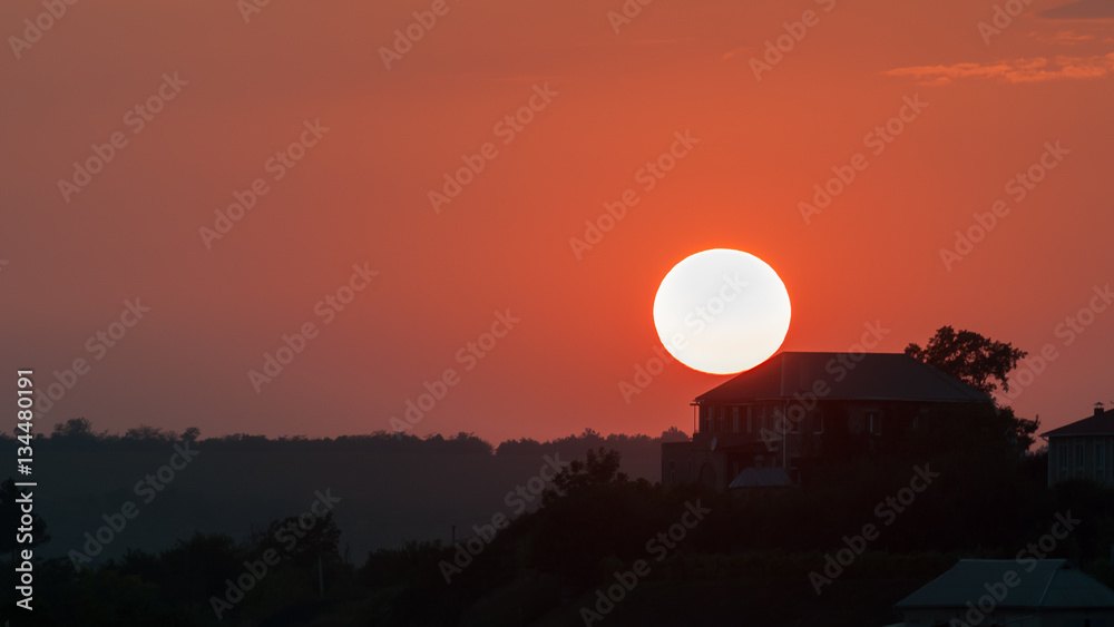 Large sun sets in the background of the rural house. Red, orange sunset, landscape.
