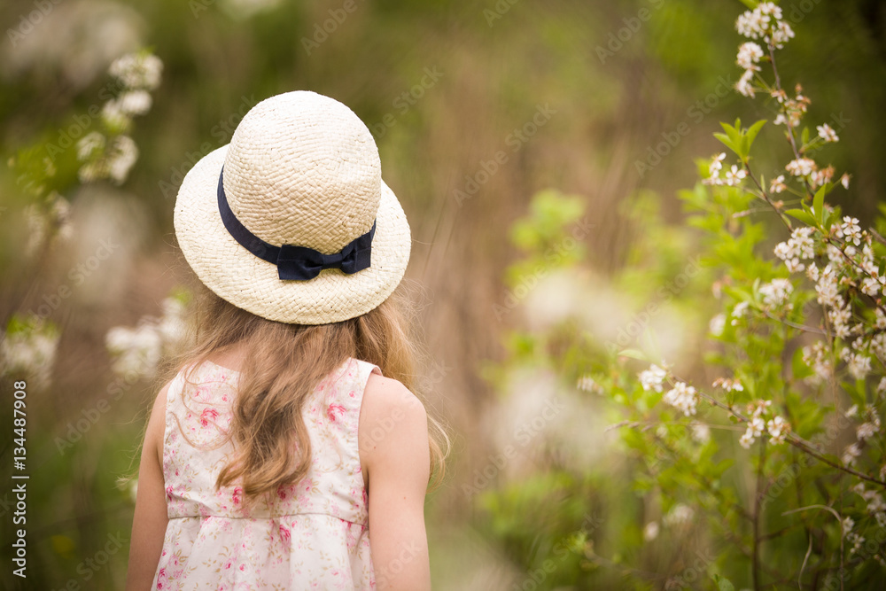 Back view on a little girl with long hair in a straw hat. Child walking in cherry blossom garden. Kid girl in a blooming park, outdoors.