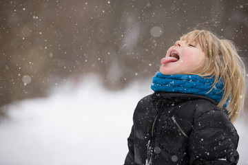Cute blond kid boy catching snowflakes with his tongue while walking in a winter park. Child having fun with snow outdoors.