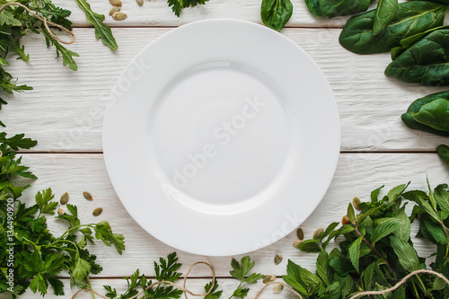 Empty white plate in seasoning frame void flat lay. Top view on blank porcelain dish bordered with fresh green  free space. Cooking  kitchen  menu  spices  cuisine concept